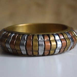 Pode incluir: Um bracelete de ouro, prata e cobre com um design texturizado e ondulado.