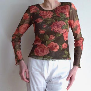 May include: A long-sleeved mesh top with a rose floral design in red and pink on a dark brown backdrop. The top has a round neck and a ruffled hem. The sleeves are long and fitted, suitable for layering.