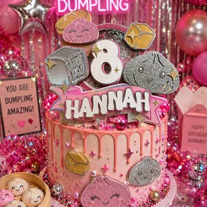 May include: A pink frosted cake with a drip design, topped with a large number 8, a banner reading HANNAH, and glittery dumpling-shaped decorations. The cake features various dumpling icons and star accents, creating a festive, themed birthday display.