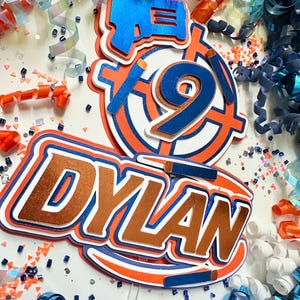 May include: A cake topper featuring the name "DYLAN" in copper letters with blue and orange accents. The topper also includes a blue number "9" inside a red and white target, and a blue toy gun.