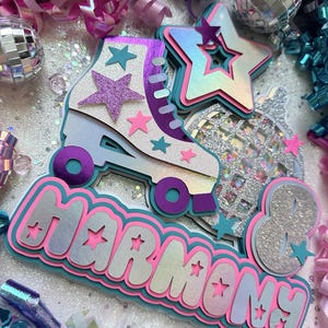 May include: A colourful party decoration with the name "Harmony" in large, glittery letters. The decoration features a roller skate, a disco ball, and stars. The colours are pink, blue, purple, and silver.