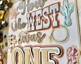 May include: A rustic-themed party sign with the text "How the West was One" in brown, white, and orange lettering. The sign features various western-themed illustrations, including a cactus, a cowboy boot, a lasso, and a gun.