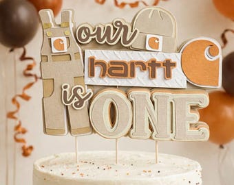 May include: A layered paper cake topper featuring a tan pair of overalls, a beanie, and the text "our hartt is ONE" in brown and white. The topper includes a circular orange logo graphic and is mounted on thin wooden sticks atop a white frosted cake.