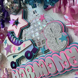 May include: A silver glitter disco ball, a pink and teal roller skate, and a pink, teal, and silver layered name banner that reads "HARMONY". The background is white with silver glitter and pink and teal streamers.