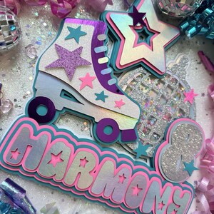 May include: A multi-layered cake topper featuring a roller skate, disco ball, and stars. The roller skate is white with purple and teal accents. The disco ball is silver with glitter. The stars are pink, white, and teal. The words "Harmony 8" are written in a pink, white, and teal font.