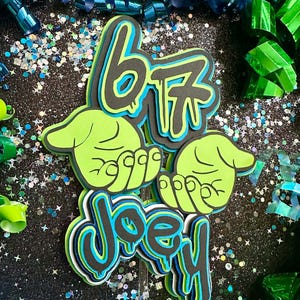 Puede incluir: Un adorno para pastel en capas con el texto "67" en negro, azul y verde, con el nombre "Joey" debajo. El adorno presenta manos verdes y está sobre un fondo negro brillante con confeti.