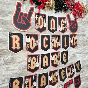 May include: A rock and roll themed banner set with black, red, and gold accents. The banner spells out "ROCK BABY" and "LAKELY" with guitar and hand sign embellishments. Decorative red, black, and gold streamers are at the top.