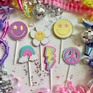Puede incluir: Una colección de coloridas decoraciones para fiestas, que incluyen adornos para pasteles con caras sonrientes, flores, arcoíris, rayos y símbolos de la paz. Rodeado de bolas de discoteca, confeti y serpentinas de colores.