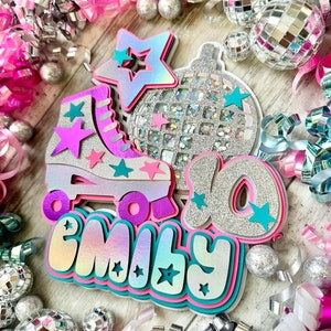 May include: A pink, teal and silver birthday cake topper featuring a roller skate, a disco ball and the name "Emily" with a star design. The topper is surrounded by silver and pink confetti.