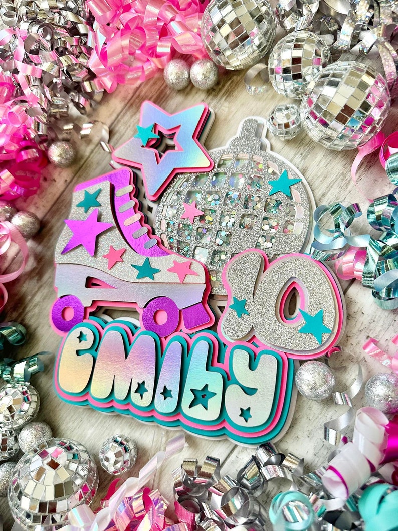 May include: A pink, teal and silver birthday cake topper featuring a roller skate, a disco ball and the name "Emily" with a star design. The topper is surrounded by silver and pink confetti.