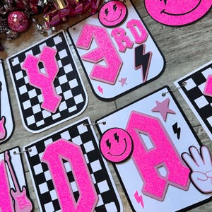 May include: A decorative banner featuring white cards with a black and white checkered border. The cards display bright pink glittery letters, smiley faces with lightning bolt eyes, pink stars, guitars, and hand signs, all strung together with silver rings.