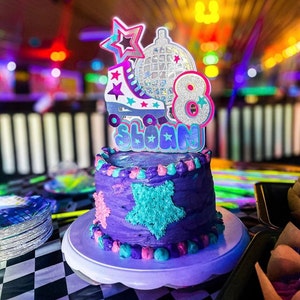 May include: Purple and blue birthday cake with a roller skate, disco ball, and the number 8 on top. The cake is decorated with stars and the name "Sloan" is written on the cake topper.