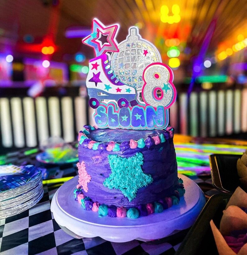 May include: Purple and blue birthday cake with a roller skate, disco ball, and the number 8 on top. The cake is decorated with stars and the name "Sloan" is written on the cake topper.