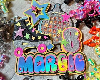 May include: A colourful birthday party decoration featuring the name "Margie" in rainbow iridescent letters, a silver roller skate with a star, a silver disco ball, and a silver number 8. The decorations are on a wooden surface.