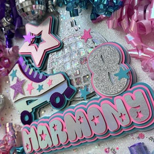 May include: A pink, blue and silver birthday cake topper with the name "Harmony" and the number "8" in a glittery font. The topper also features a roller skate, a disco ball and stars.