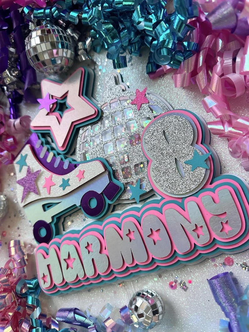 May include: A pink, blue and silver birthday cake topper with the name "Harmony" and the number "8" in a glittery font. The topper also features a roller skate, a disco ball and stars.
