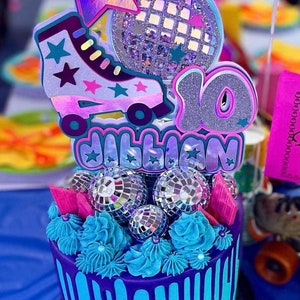 May include: A purple and blue cake decorated with a roller skate, disco ball, and the number 10. The cake is topped with blue frosting and silver disco balls. The cake is decorated with the name "Jillian".