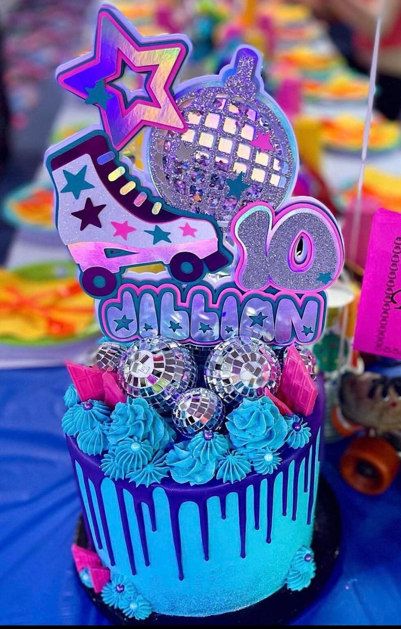 May include: A purple and blue cake decorated with a roller skate, disco ball, and the number 10. The cake is topped with blue frosting and silver disco balls. The cake is decorated with the name "Jillian".