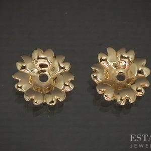May include: Two gold-coloured, flower-shaped earring backs. Each back has a central hole and petal-like edges. The jewellery pieces are displayed on a dark surface. The image is well-lit, highlighting the details of the metalwork.