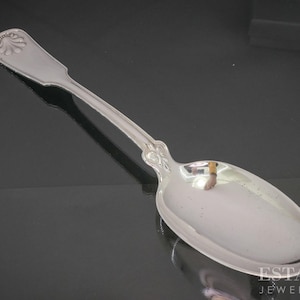 May include: A silver spoon with a decorative shell design on the handle. The spoon has a long, slender handle and a rounded bowl. The spoon is highly polished and reflects the surrounding environment. The spoon is likely for serving.
