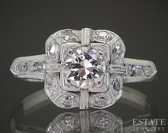 Può includere: Un anello in platino con un grande diamante centrale a taglio rotondo. L'anello presenta un'aureola quadrata di diamanti più piccoli e diamanti aggiuntivi incastonati lungo la fascia. L'anello è posto su uno sfondo scuro.