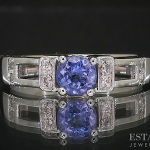 May include: A silver ring featuring a central, round, blue gemstone, with smaller diamonds on either side. The ring has a contemporary design with rectangular cutouts. The ring is shown on a reflective black surface.