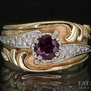 May include: A gold and silver ring with a large, round, deep red gemstone at the center. The ring has an ornate design with swirling gold accents and silver filigree details. The ring is set against a black background.