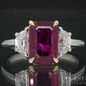 May include: A platinum ring featuring a large, rectangular, deep red ruby center stone, flanked by two smaller, white, rectangular diamonds. The ruby is accented with gold prongs. The ring is set against a black background.