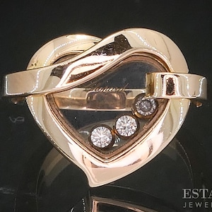 May include: A gold-toned heart-shaped pendant with a clear center, featuring three diamonds. The pendant has a unique design with a curved top and a horizontal bar across the center. The words "Chopard" are visible inside the clear center.