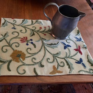 May include: A square cream-colored fabric mat featuring an intricate embroidered pattern of swirling green vines, leaves, and multi-colored flowers in shades of red, blue, and gold. A metallic silver-toned pitcher with a handle sits on the mat.