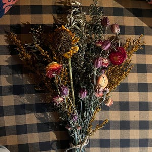 May include: A bouquet of dried flowers in shades of brown, yellow, red, and purple. The flowers are tied together with twine and are arranged on a brown and black checkered surface.