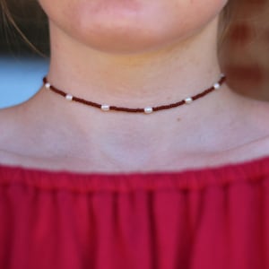 May include: A close-up of a brown beaded choker necklace with white pearl accents. The necklace is worn by a person with a red off-the-shoulder top.