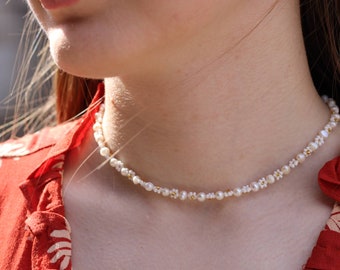 May include: A delicate pearl and gold choker necklace with small flower accents. The necklace is worn by a person with long brown hair and a red floral shirt.