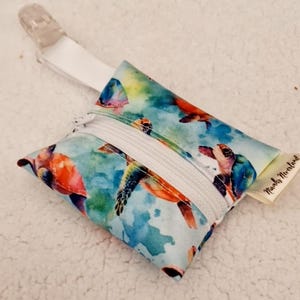 Op de afbeelding: Een witte ritszak met een clip om aan een tas of kinderwagen te bevestigen. De zak is versierd met een aquarelpatroon van zeeschildpadden die in de oceaan zwemmen. De tekst "Naughty Nerdland" staat gedrukt op een label dat aan de zak is bevestigd.