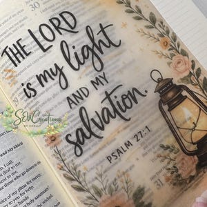 Peut inclure: Page de la Bible avec l'inscription "THE LORD is my light AND MY salvation" en écriture noire. Le design comprend des éléments floraux et une illustration de lanterne. Le Psaume 27:1 est également visible.
