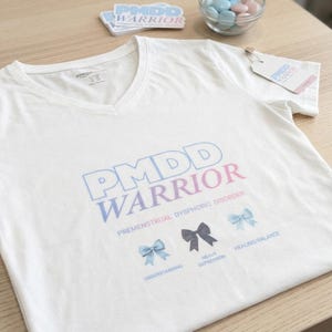 May include: A white V-neck t-shirt featuring the text PMDD WARRIOR and PREMENSTRUAL DYSPHORIC DISORDER in blue and pink. Below the text are three bows in light blue, dark gray, and light blue with the labels UNDERSTANDING, HEAVY DEPRESSION, and HEALING/BALANCE.