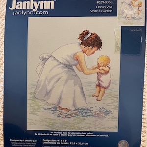 May include: A Janlynn counted cross stitch kit titled Ocean Visit, featuring a design of a person in a white dress holding a baby. The packaging includes the design size of 9 inches by 12 inches, 14-count cotton Aida fabric, 6-strand cotton floss, a needle, a graph, floss cards, and instructions.