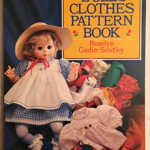 May include: A book titled "Dolls' Clothes Pattern Book" by Roselyn Gadia-Smitley. The cover features a doll wearing a white and blue gingham dress and a straw hat. The doll is surrounded by fabric scraps, sewing supplies, and a red pincushion.
