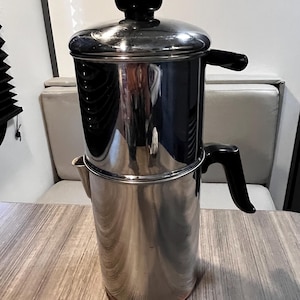 May include: A stainless steel coffee percolator with a black handle and lid knob. The percolator sits on a wooden table. The top of the percolator is a shiny, reflective metal, and the bottom has a copper-colored base.
