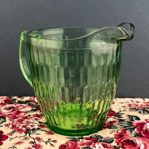 May include: A green glass pitcher with a handle and a pouring spout. The pitcher has a textured, geometric pattern. The pitcher is sitting on a floral patterned tablecloth with red and pink roses.