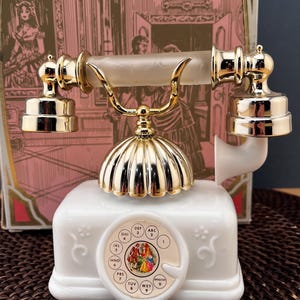 May include: A white rotary telephone with gold-tone metallic accents and a frosted glass handle. The base features a circular dial with a central illustration of two figures in period clothing, surrounded by standard alphanumeric keypad markings.