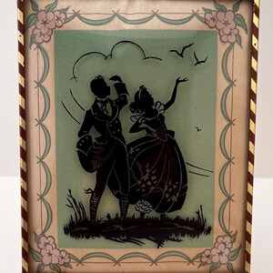 May include: A rectangular framed silhouette art piece featuring a black cutout of a couple in period clothing on a light green background. The frame has a red and gold striped border with floral corner accents. The couple stands on dark grass.