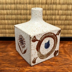 May include: A square ceramic vase with a speckled white finish and a narrow neck. The vase is decorated with brown and blue abstract floral designs. The vase sits on a wooden surface.