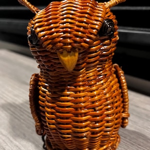 May include: A brown woven wicker owl figurine featuring a rounded body, pointed ear tufts, and a small triangular beak. The owl has dark, circular eyes and a detailed, textured surface created by the intricate interlacing of the brown wicker material.