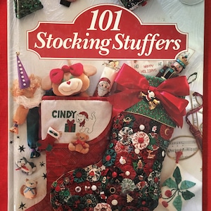 May include: A book titled "101 Stocking Stuffers" with a red and white stocking filled with various Christmas-themed items, including a stuffed animal, a small toy, and a bow. The stocking is decorated with red, green, and white fabric flowers.