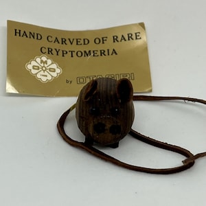 May include: Hand carved wooden pig figurine with a brown leather cord. The tag reads "Hand Carved of Rare Cryptomeria by Otagirl".