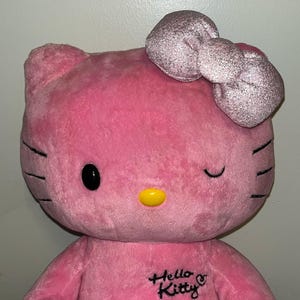Puede incluir: Peluche rosa Hello Kitty con un lazo de purpurina plateada. El juguete tiene ojos negros, una nariz amarilla y el texto "Hello Kitty" bordado en su pecho.
