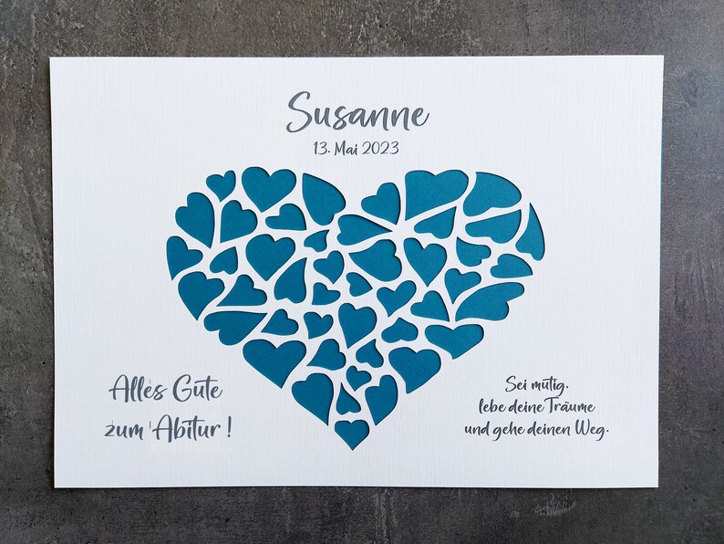 May include: A white card with a teal heart-shaped design filled with smaller hearts. The name "Susanne" and the date "13. Mai 2023" are printed at the top. Text in German is also included.