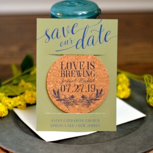 May include: A wedding save-the-date card with a cork coaster. The card is olive green with the words "save our date" in blue script. The coaster reads "Love is Brewing" with a date and names.