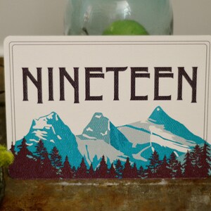 May include: A white card with the word "NINETEEN" printed in black. The card features a graphic of three blue mountains with snow-capped peaks and a forest of dark purple trees in the foreground.